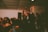 A live music performance is taking place in a compact indoor venue with a crowd of people gathered around. The lead singer is passionately holding a microphone and singing, surrounded by audience members who appear engaged and energetic. The setting is dimly lit, creating an intimate ambiance.