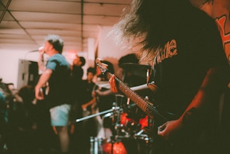 A band is performing in a small, dimly-lit venue, with one person singing into a microphone and another playing an electric guitar. The atmosphere is energetic, with blurred motion capturing the intensity of the performance. The audience is partially visible, indicating a crowded space.