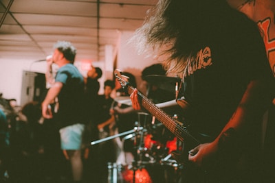 A band is performing in a small, dimly-lit venue, with one person singing into a microphone and another playing an electric guitar. The atmosphere is energetic, with blurred motion capturing the intensity of the performance. The audience is partially visible, indicating a crowded space.