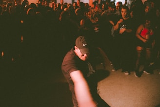 A person wearing a cap and dark clothing is energetically dancing in a dimly lit room filled with a crowd. The crowd watches attentively, with some capturing the moment or cheering. The lighting creates dramatic shadows, enhancing the dynamic and vibrant atmosphere.