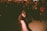 A person wearing a cap and dark clothing is energetically dancing in a dimly lit room filled with a crowd. The crowd watches attentively, with some capturing the moment or cheering. The lighting creates dramatic shadows, enhancing the dynamic and vibrant atmosphere.
