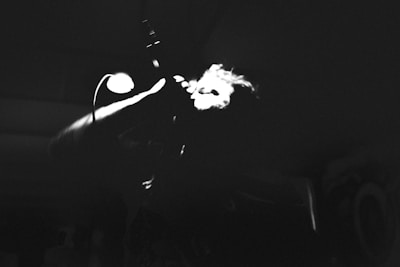 A high contrast black and white image depicts a person singing passionately into a microphone with dramatic lighting highlighting their face and hand. The background is dark, emphasizing the illuminated areas.
