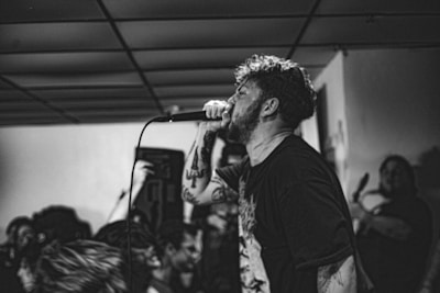 A black and white image capturing a passionate singer performing in a small, crowded venue. The singer holds a microphone close to their mouth and appears to be deeply immersed in their performance. The background shows a few audience members and a ceiling with a grid pattern.