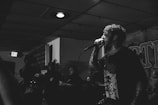 A black and white image featuring a vocalist passionately performing on stage with a microphone. The setting appears to be an indoor venue with a small crowd in the background. The performer has tattoos and is wearing a dark shirt with graphic art. The ambiance suggests a dimly lit environment with a low ceiling and visible exit sign.