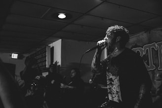 A black and white image featuring a vocalist passionately performing on stage with a microphone. The setting appears to be an indoor venue with a small crowd in the background. The performer has tattoos and is wearing a dark shirt with graphic art. The ambiance suggests a dimly lit environment with a low ceiling and visible exit sign.