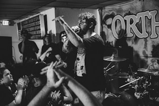 A black and white photograph of a live music performance in a small venue. A man with tattoos and facial hair is energetically performing, holding a microphone with one hand and gesturing with the other. The background features a drum set, amplifiers, and a banner with large lettering. The audience is close to the stage, with raised hands and expressions of engagement.
