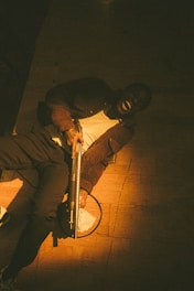 A person is lying on the floor in dim lighting, holding a shotgun across their body. There is a stack of money placed beside them, partially illuminated by a warm, focused light coming from above.