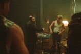 A group of men are gathered around a table inside a dimly lit room. The table is cluttered with playing cards, cash, and some other objects. One man in a suit appears to be speaking or arguing with others, while another man, wearing a white tank top and sunglasses, stands with a stern expression. The background features dark wooden furniture, a lampshades casting a warm light, and a generally smoky atmosphere.