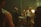 A group of men are gathered around a table inside a dimly lit room. The table is cluttered with playing cards, cash, and some other objects. One man in a suit appears to be speaking or arguing with others, while another man, wearing a white tank top and sunglasses, stands with a stern expression. The background features dark wooden furniture, a lampshades casting a warm light, and a generally smoky atmosphere.