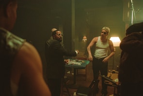 A group of men are gathered around a table inside a dimly lit room. The table is cluttered with playing cards, cash, and some other objects. One man in a suit appears to be speaking or arguing with others, while another man, wearing a white tank top and sunglasses, stands with a stern expression. The background features dark wooden furniture, a lampshades casting a warm light, and a generally smoky atmosphere.