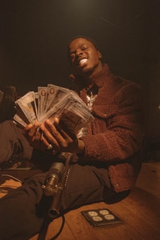 A person dressed in a brown jacket is holding a large fan of cash in their hands. The lighting creates a dramatic and intense atmosphere, casting shadows and emphasizing textures. The setting includes additional objects like a gun resting on their lap, giving a sense of edginess.