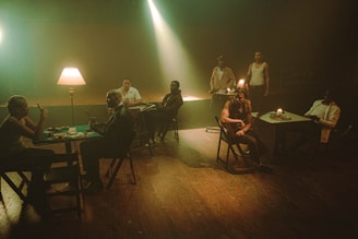 Several people sit at tables in a dimly lit room with a warm, moody atmosphere. A lamp and candle provide soft lighting, casting shadows across their faces. Cards and chips are on the tables, suggesting a game of poker. The setting is rustic, with wooden floors and simple furniture.