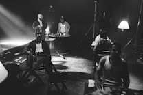 A black and white scene with several people gathered around tables, illuminated by dim lamps. Bottles and drinks are visible, and one person appears to be dealing cards. The setting appears relaxed and casual, with a retro vibe. The presence of stage lights and a visible exit sign suggests a staged or theatrical atmosphere.