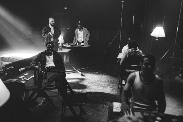 A black and white scene with several people gathered around tables, illuminated by dim lamps. Bottles and drinks are visible, and one person appears to be dealing cards. The setting appears relaxed and casual, with a retro vibe. The presence of stage lights and a visible exit sign suggests a staged or theatrical atmosphere.