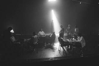 A dimly lit room with several people gathered around tables. The setting appears to be tense, with one person standing in a spotlight, while others are seated or standing, engaged in what seems like a serious conversation. The atmosphere is reminiscent of a noir film or a detective scene.
