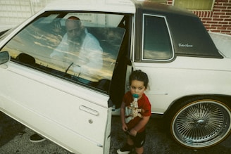 A young child holding a giraffe toy stands beside a white vintage car with the driver-side door open. A man is seen sitting inside the car, gazing outward through the window. The car is parked near a brick wall and has distinctive, wire-spoke wheels.