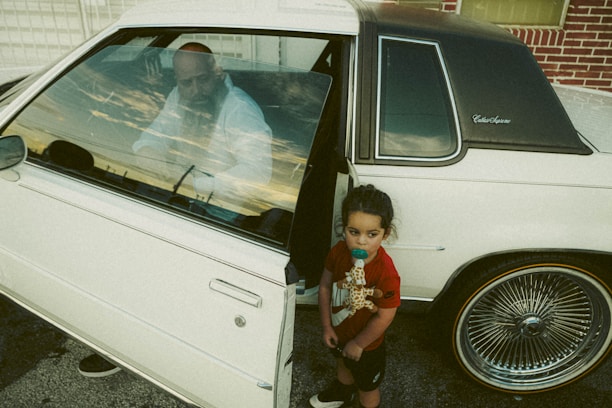A young child holding a giraffe toy stands beside a white vintage car with the driver-side door open. A man is seen sitting inside the car, gazing outward through the window. The car is parked near a brick wall and has distinctive, wire-spoke wheels.