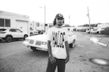 A person wearing a T-shirt with a bold design and a cap stands in front of a classic car in an industrial area. There are several parked cars and buildings with garage-style doors in the background. The ground is wet, with puddles reflecting the surroundings.