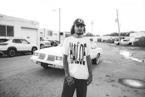 A person wearing a T-shirt with a bold design and a cap stands in front of a classic car in an industrial area. There are several parked cars and buildings with garage-style doors in the background. The ground is wet, with puddles reflecting the surroundings.