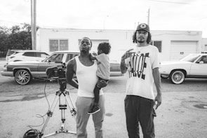 Two individuals stand in a parking lot beside a professional camera setup on a tripod. One of them is holding a child. Behind them, several cars are parked, including vintage models. The setting appears casual, and the image is in black and white.