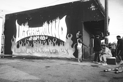 A black and white image featuring a mural on a building wall with the words 'Underground - Ult Don't Fold' surrounded by flame graphics. Several people are gathered near the wall. One person sits on a cooler, while others stand nearby. A child is seen perched on another person's shoulders.
