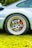 A close-up view of a car wheel featuring a polished alloy rim with a distinctive design. The wheel has large perforations and a visible car logo at the center. There is a bright red brake caliper behind the rim, adding contrast to the metallic finish. The car's sleek bodywork is visible, painted in a light color, and the vehicle is parked on well-maintained green grass.