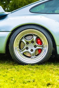A close-up view of a car wheel featuring a polished alloy rim with a distinctive design. The wheel has large perforations and a visible car logo at the center. There is a bright red brake caliper behind the rim, adding contrast to the metallic finish. The car's sleek bodywork is visible, painted in a light color, and the vehicle is parked on well-maintained green grass.