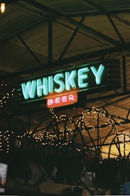 Neon signs displaying the words 'WHISKEY' and 'BEER' are framed against a backdrop of illuminated string lights, creating a rustic and cozy bar atmosphere. The structure appears to be an indoor space, possibly under a wooden ceiling with metal beams.