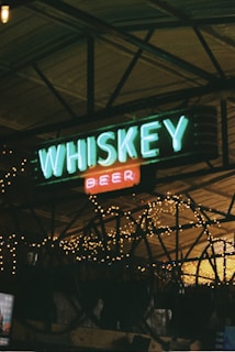 Neon signs displaying the words 'WHISKEY' and 'BEER' are framed against a backdrop of illuminated string lights, creating a rustic and cozy bar atmosphere. The structure appears to be an indoor space, possibly under a wooden ceiling with metal beams.