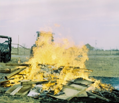 A large fire consumes a pile of wooden debris on an open field. In the background, a vehicle and a tractor are partially visible, with a faint horizon line and sparse, dry vegetation.
