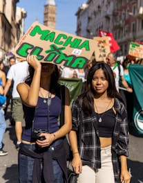 Two young women are walking in a street protest or march, holding a sign that reads 'MACHA CLIMA' with green letters. The background shows more people, likely participating in the same protest, holding various signs. The setting is an urban street lined with buildings.