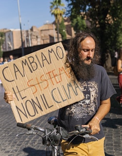 A man with long hair and a beard stands outside holding a cardboard protest sign that reads 'CAMBIAMO IL SISTEMA NON IL CLIMA' in Italian. He is next to a bicycle and wears a dark t-shirt and yellow pants. The urban background shows a palm tree and out-of-focus buildings.