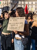 A group of young people are gathered in a city square holding signs with messages written in Italian. The scene is lively and appears to be a protest or demonstration, with several individuals engaging in conversation. The buildings in the background suggest an urban setting, and some participants are casually dressed, suggesting a warm or mild day.