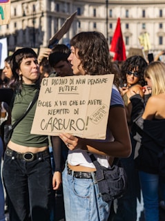 A group of young people are gathered in a city square holding signs with messages written in Italian. The scene is lively and appears to be a protest or demonstration, with several individuals engaging in conversation. The buildings in the background suggest an urban setting, and some participants are casually dressed, suggesting a warm or mild day.