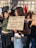 A group of young people are gathered in a city square holding signs with messages written in Italian. The scene is lively and appears to be a protest or demonstration, with several individuals engaging in conversation. The buildings in the background suggest an urban setting, and some participants are casually dressed, suggesting a warm or mild day.
