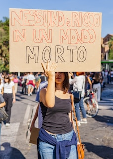 A person is holding a protest sign with bold, hand-written Italian text in a crowded outdoor setting. The scene suggests an organized march or demonstration with several people in the background, some walking and others standing, while wearing casual clothing.