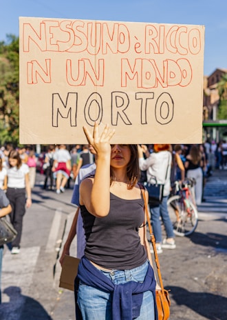 A person is holding a protest sign with bold, hand-written Italian text in a crowded outdoor setting. The scene suggests an organized march or demonstration with several people in the background, some walking and others standing, while wearing casual clothing.