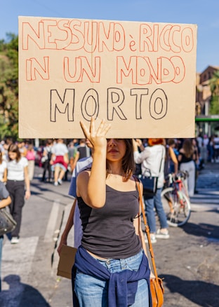 A person is holding a protest sign with bold, hand-written Italian text in a crowded outdoor setting. The scene suggests an organized march or demonstration with several people in the background, some walking and others standing, while wearing casual clothing.