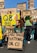 Several people are gathered outdoors, holding various signs or placards. Some signs display the names of different oil companies such as 'Q8 Oils,' 'ABU DHABI NATIONAL OIL CO,' 'Nigerian National Petroleum Corp,' 'Sonatrach SPA,' and others. One man with long hair tied in a ponytail is kneeling and holding a sign. The scene appears to be part of a protest or demonstration, with an old tower structure visible in the background.