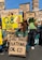 Several people are gathered outdoors, holding various signs or placards. Some signs display the names of different oil companies such as 'Q8 Oils,' 'ABU DHABI NATIONAL OIL CO,' 'Nigerian National Petroleum Corp,' 'Sonatrach SPA,' and others. One man with long hair tied in a ponytail is kneeling and holding a sign. The scene appears to be part of a protest or demonstration, with an old tower structure visible in the background.