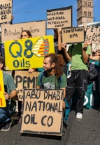 Several people are gathered outdoors, holding various signs or placards. Some signs display the names of different oil companies such as 'Q8 Oils,' 'ABU DHABI NATIONAL OIL CO,' 'Nigerian National Petroleum Corp,' 'Sonatrach SPA,' and others. One man with long hair tied in a ponytail is kneeling and holding a sign. The scene appears to be part of a protest or demonstration, with an old tower structure visible in the background.