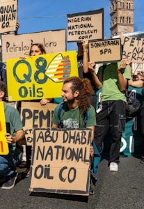 Several people are gathered outdoors, holding various signs or placards. Some signs display the names of different oil companies such as 'Q8 Oils,' 'ABU DHABI NATIONAL OIL CO,' 'Nigerian National Petroleum Corp,' 'Sonatrach SPA,' and others. One man with long hair tied in a ponytail is kneeling and holding a sign. The scene appears to be part of a protest or demonstration, with an old tower structure visible in the background.