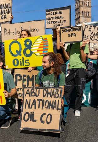 Several people are gathered outdoors, holding various signs or placards. Some signs display the names of different oil companies such as 'Q8 Oils,' 'ABU DHABI NATIONAL OIL CO,' 'Nigerian National Petroleum Corp,' 'Sonatrach SPA,' and others. One man with long hair tied in a ponytail is kneeling and holding a sign. The scene appears to be part of a protest or demonstration, with an old tower structure visible in the background.