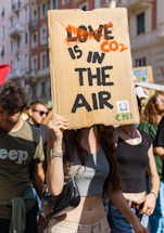 A group of people are participating in a protest or demonstration. One individual in the foreground holds a cardboard sign with the words 'CO2 IS IN THE AIR', which is creatively edited from the phrase 'LOVE IS IN THE AIR'. The scene is outdoors, likely in an urban setting with buildings visible in the background. The atmosphere appears lively and crowded.