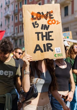 A group of people are participating in a protest or demonstration. One individual in the foreground holds a cardboard sign with the words 'CO2 IS IN THE AIR', which is creatively edited from the phrase 'LOVE IS IN THE AIR'. The scene is outdoors, likely in an urban setting with buildings visible in the background. The atmosphere appears lively and crowded.