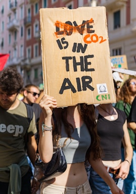 A group of people are participating in a protest or demonstration. One individual in the foreground holds a cardboard sign with the words 'CO2 IS IN THE AIR', which is creatively edited from the phrase 'LOVE IS IN THE AIR'. The scene is outdoors, likely in an urban setting with buildings visible in the background. The atmosphere appears lively and crowded.