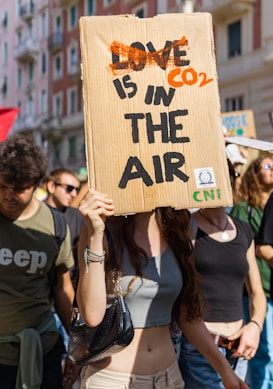 A group of people are participating in a protest or demonstration. One individual in the foreground holds a cardboard sign with the words 'CO2 IS IN THE AIR', which is creatively edited from the phrase 'LOVE IS IN THE AIR'. The scene is outdoors, likely in an urban setting with buildings visible in the background. The atmosphere appears lively and crowded.