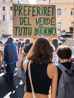 A person holding a cardboard sign with green handwritten text reading 'PREFERIREI DEL VERDE TUTTO INTORNO' among a crowd of people participating in a street protest or rally. The scene is set in an urban environment with old buildings visible in the background.