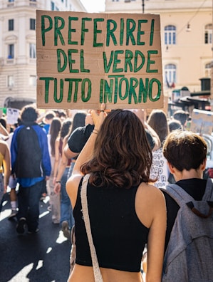 A person holding a cardboard sign with green handwritten text reading 'PREFERIREI DEL VERDE TUTTO INTORNO' among a crowd of people participating in a street protest or rally. The scene is set in an urban environment with old buildings visible in the background.