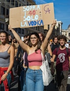 A group of young people participating in a protest or demonstration, walking down a street in a city setting with buildings in the background. The central figure, a woman, is holding up a cardboard sign with colorful text in Italian. The crowd is casually dressed, and the atmosphere suggests a peaceful and energetic gathering.